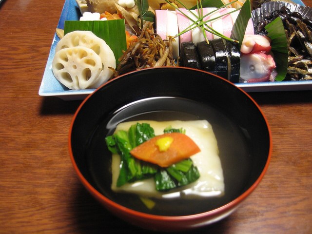 黒い椀のすまし汁に角餅と青菜、にんじんを入れたお雑煮と、奥に並ぶおせち料理